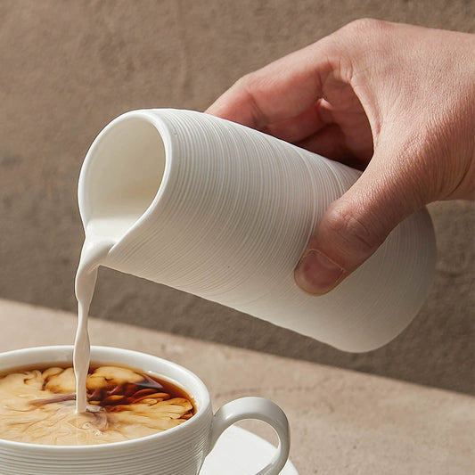 Hand pouring milk from a white pitcher into a cup of coffee on a neutral background