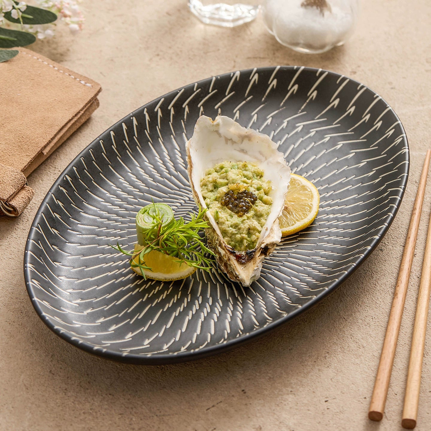 Oyster with avocado on a decorative black plate on a beige surface with chopsticks.