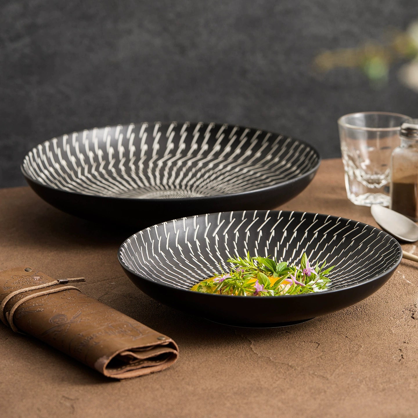 Two black ceramic bowls on a brown surface with a glass of water and a salt grinder in the background.
