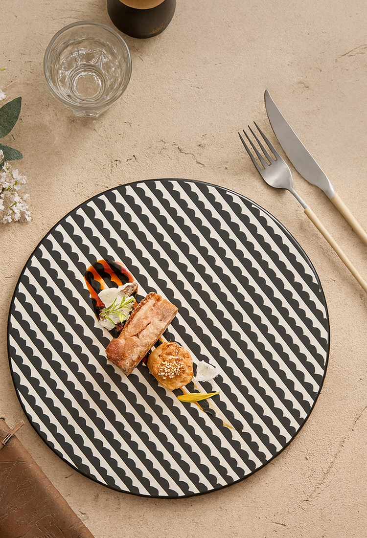 Round black and white patterned plate with a meal on a beige surface with a glass and cutlery.