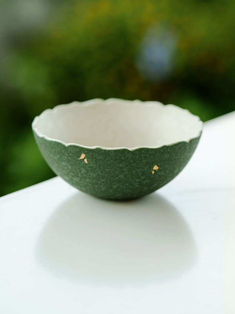 Green ceramic tea cup with white interior on a white surface with a blurred green 
