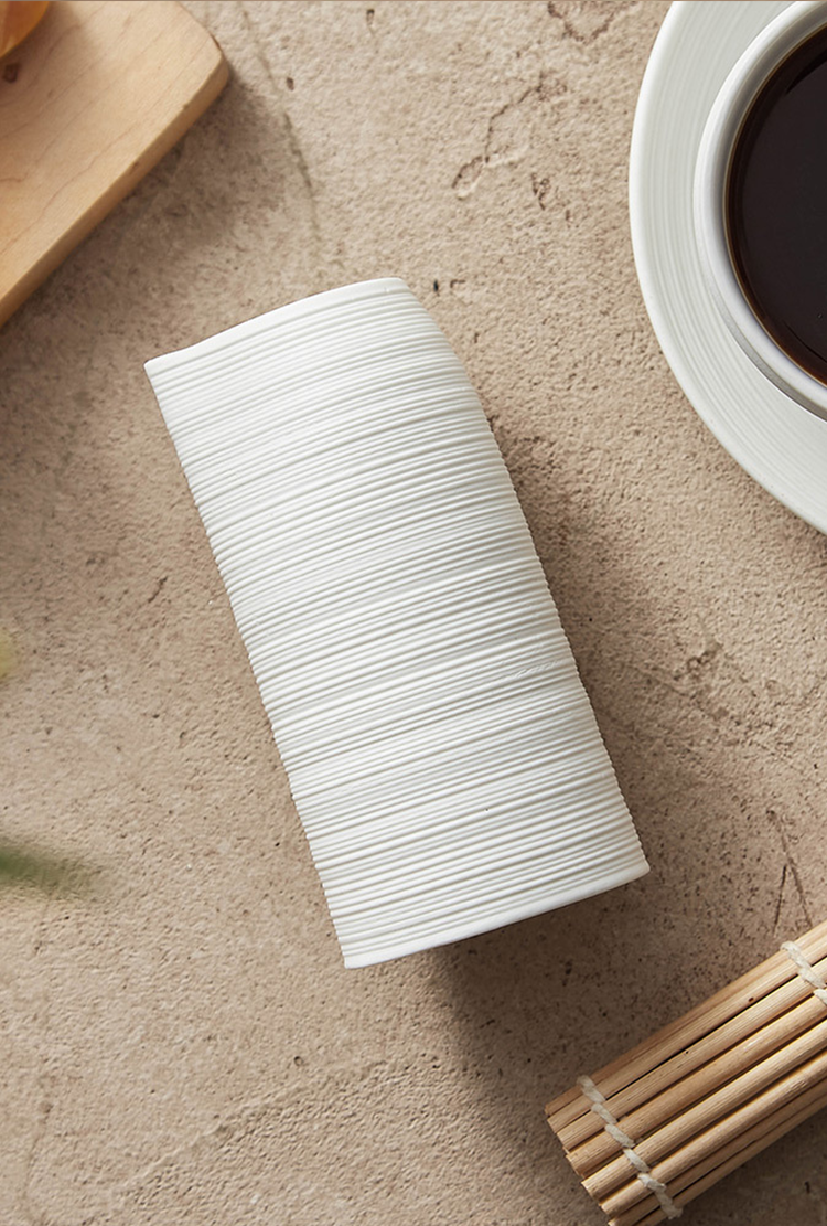 White ceramic vase on a textured surface with a cup of coffee and wooden sticks in the background.