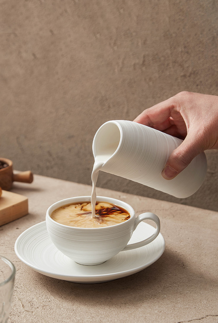 Person pouring milk from a white pitcher into a cup of coffee on a wooden table.