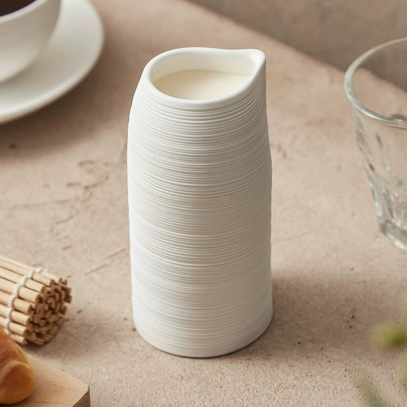 White ceramic pitcher on a beige surface with a glass and wooden items in the background
