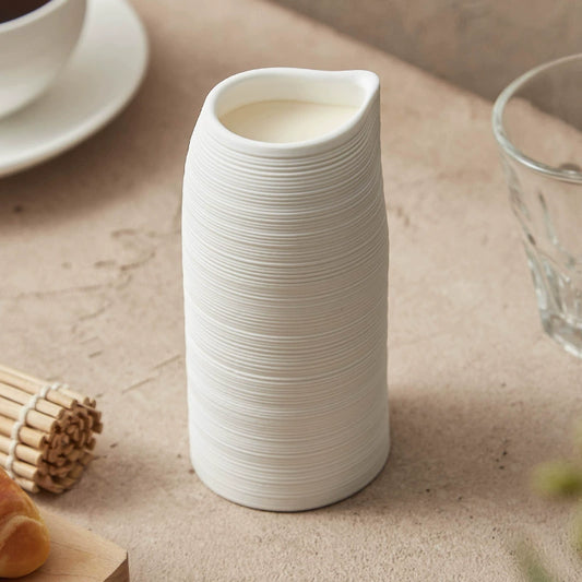 White ceramic pitcher on a beige surface with a glass and wooden items in the background