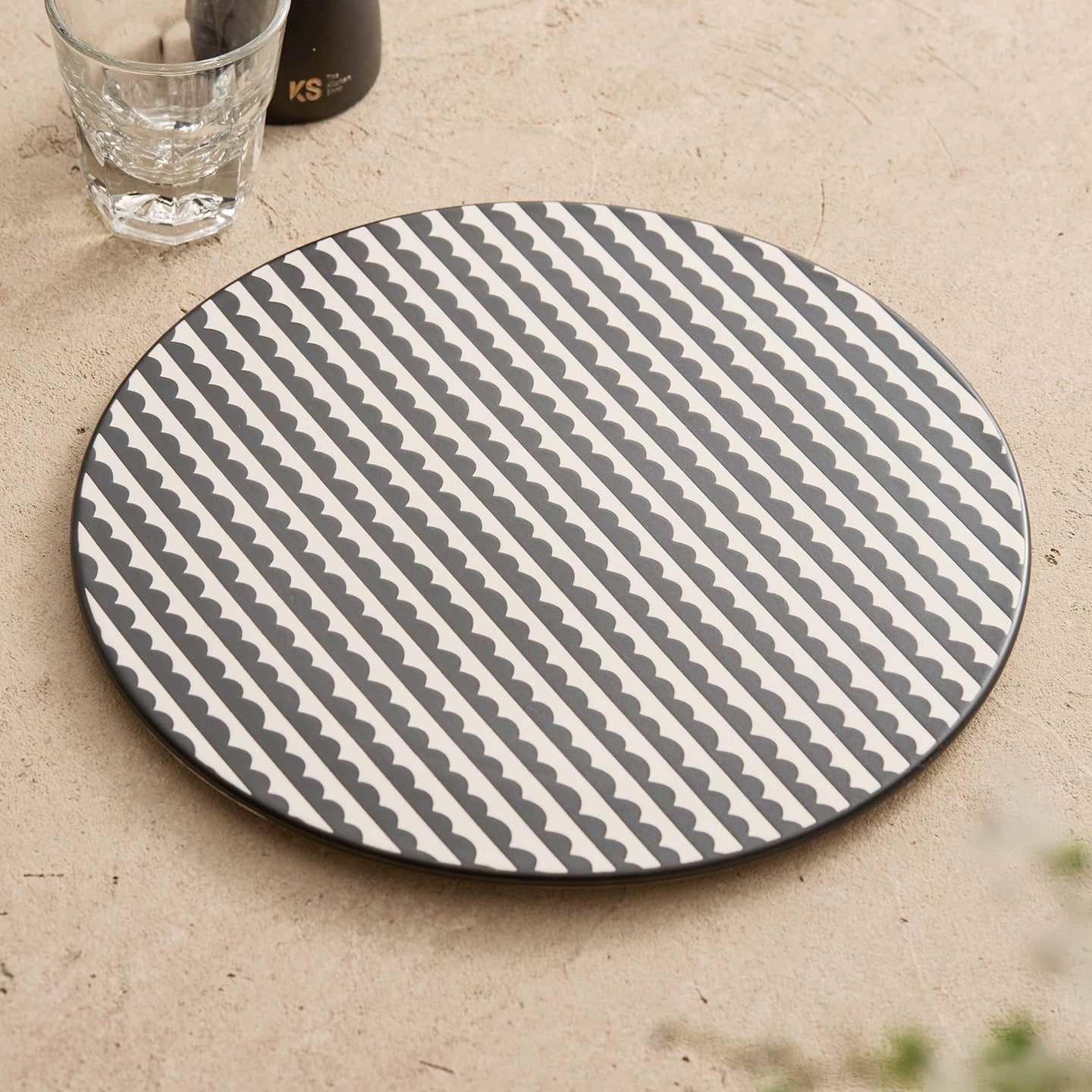 Round gray and white patterned placemat on a beige surface with a glass of water and black container.