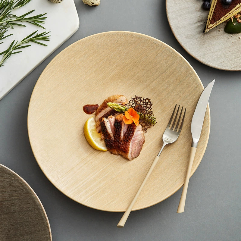 Beige plate with a dish of meat, lemon, and garnishes on a gray surface with a fork and knife.