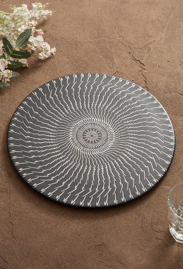 Round ceramic plate with a decorative pattern on a brown surface with flowers and a glass.