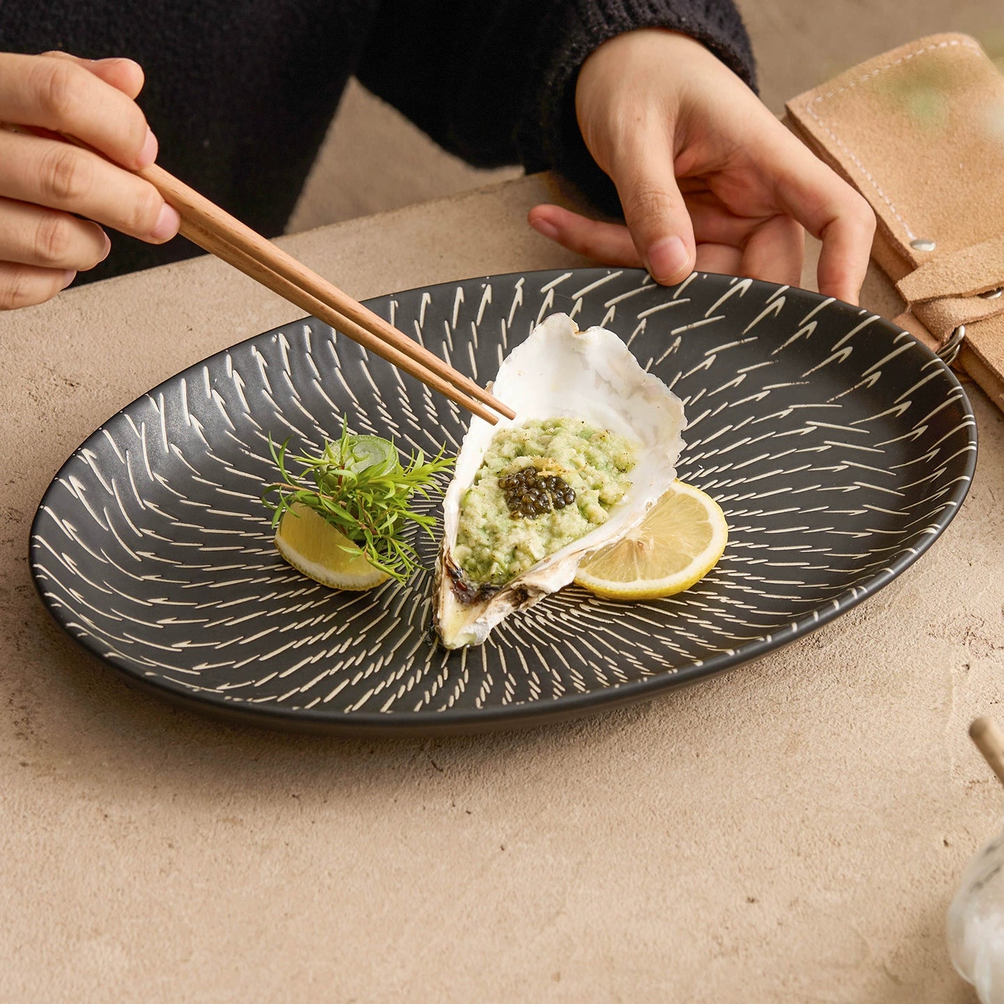 Person using chopsticks to pick up an oyster on a black decorative plate with a lemon wedge and garnish.