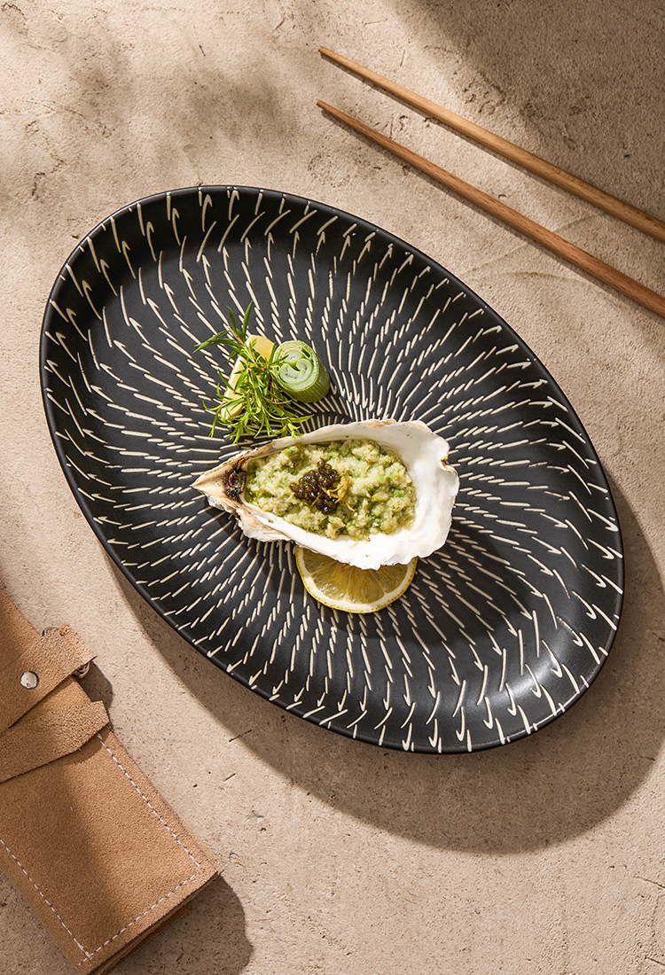 Black decorative plate with an oyster dish on a textured surface with chopsticks.